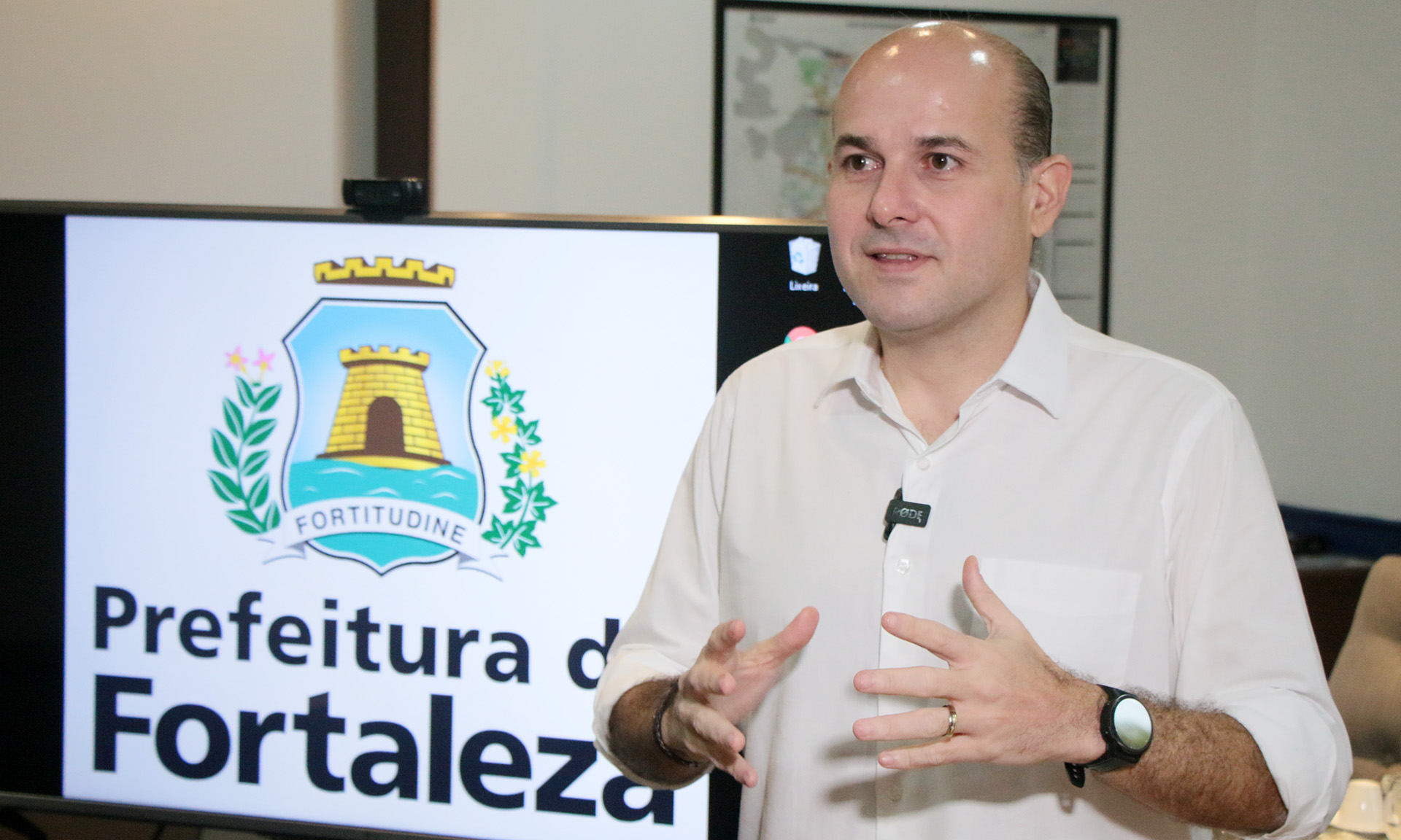 prefeito de lado falando com monitor com logo da prefeitura ao fundo